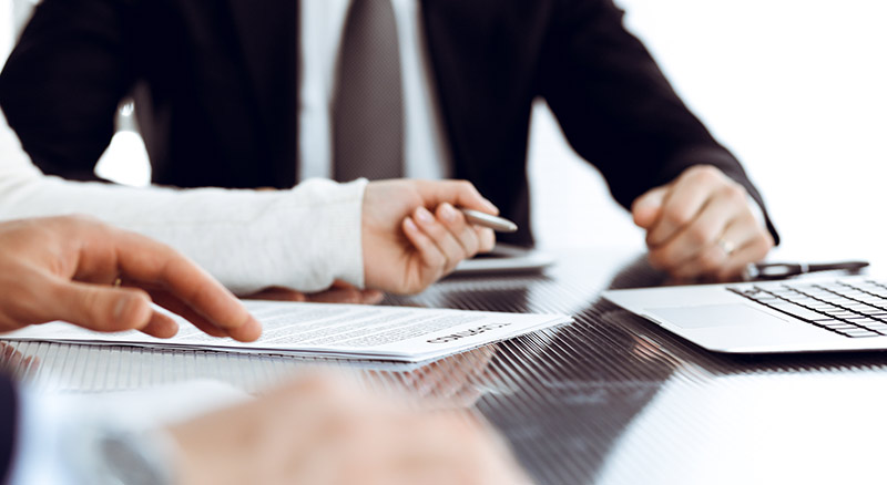 A person in a business suit is reviewing and signing documents at a desk, with a laptop partially visible to the side