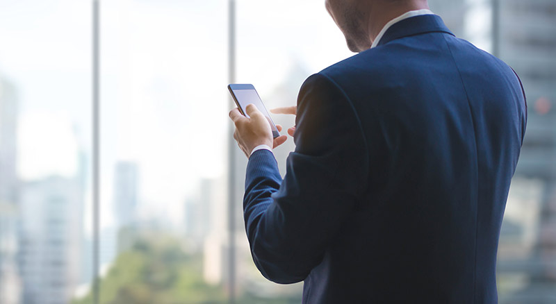 A person in a blue suit is using a smartphone by a window with a cityscape view in the background.

