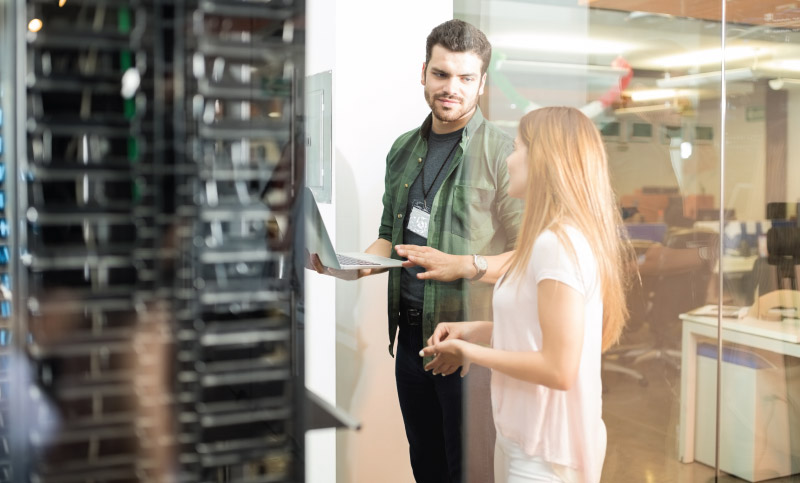Two people are in a server room, one holding a laptop and gesturing towards it while the other observes