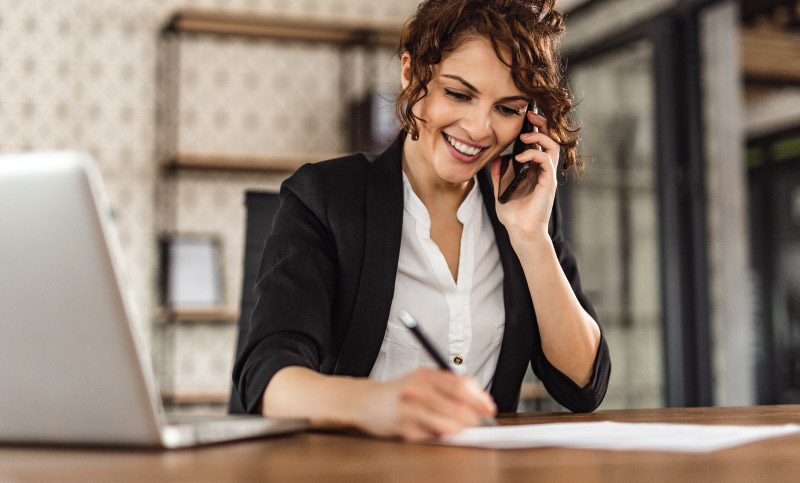 A person in business attire writes at a desk while holding a phone to their ear