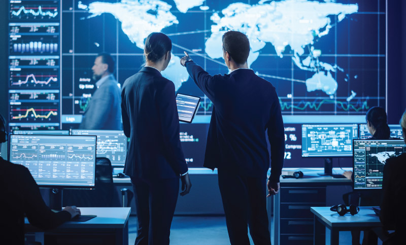 Two professionals analyze data on multiple computer screens with global maps and graphs