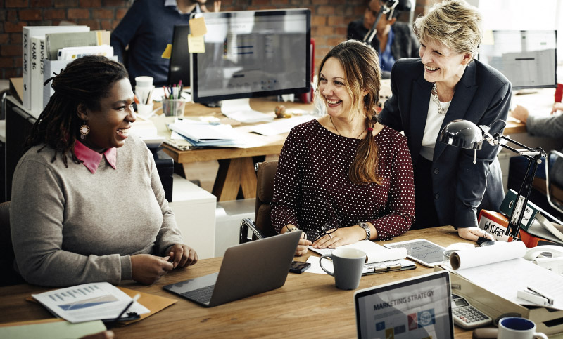 Three women are engaged in a meeting around a table cluttered with laptops and papers in an office setting